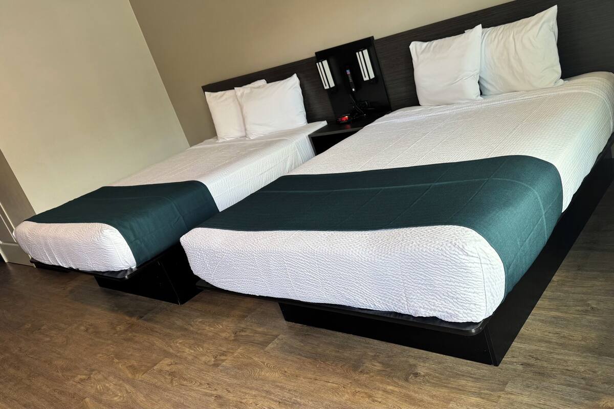 Una cama doble con ropa de cama blanca y verde agua, una mesita de noche con un reloj, y suelo de madera.