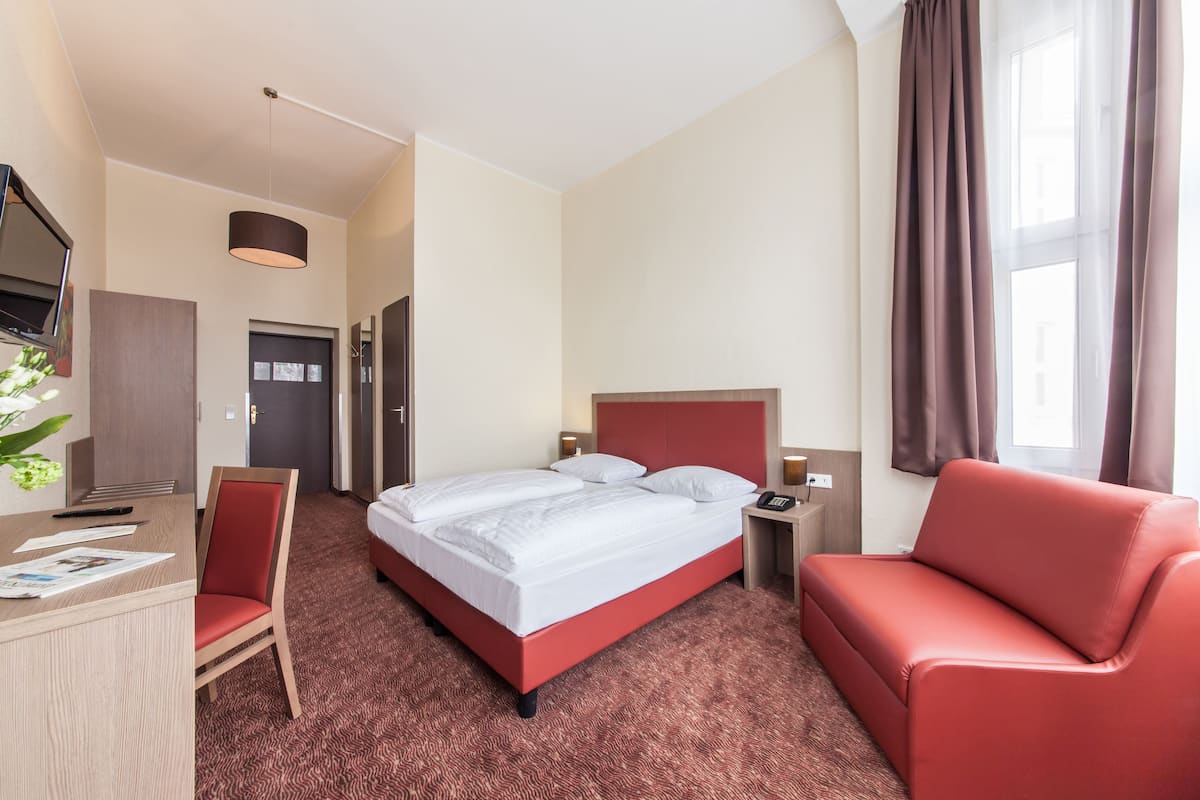 Una habitación de hotel con cama, escritorio, silla, sofá, televisor y ventana con cortinas.