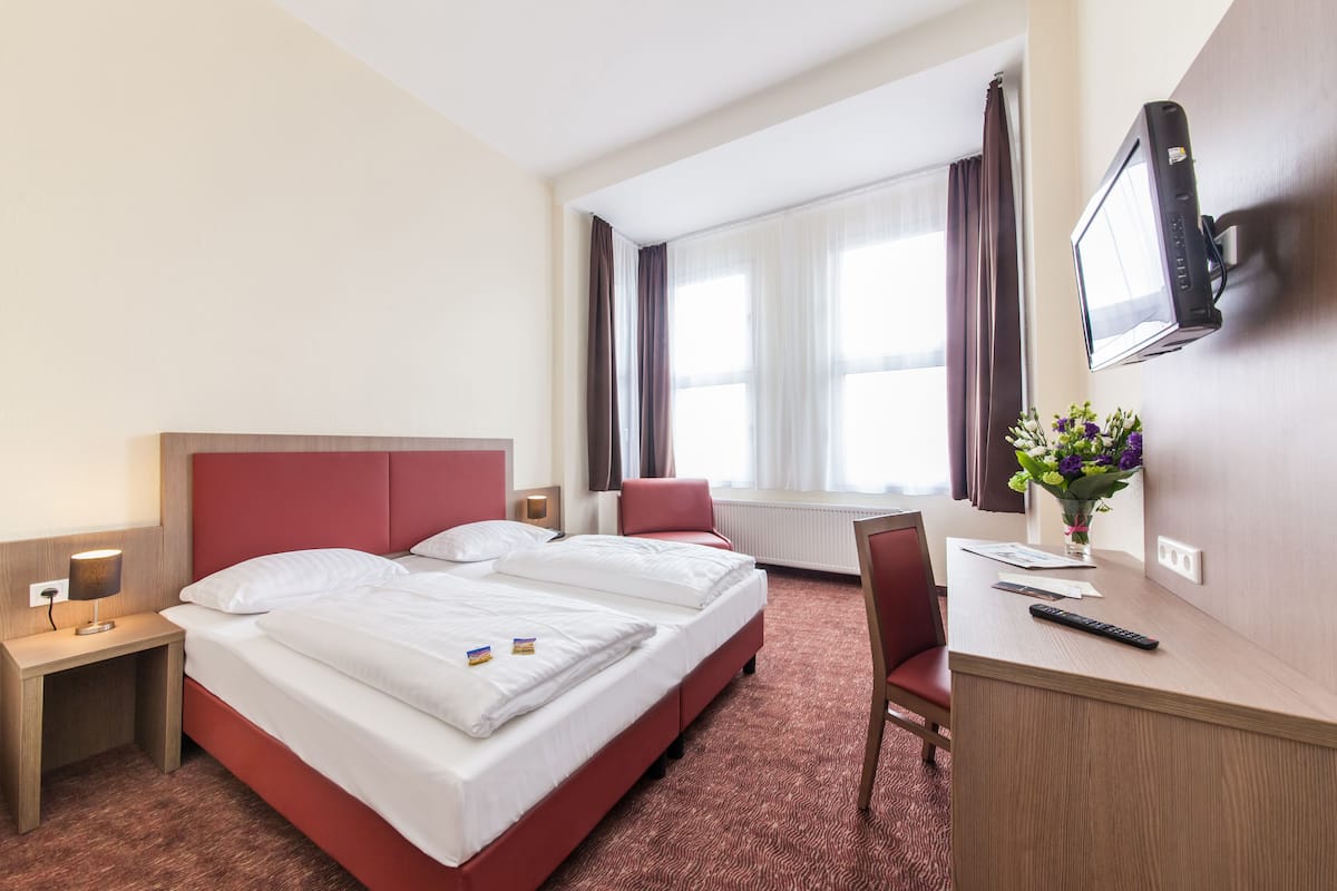 Una habitación de hotel con cama, escritorio, silla, televisión y ventana con cortinas.