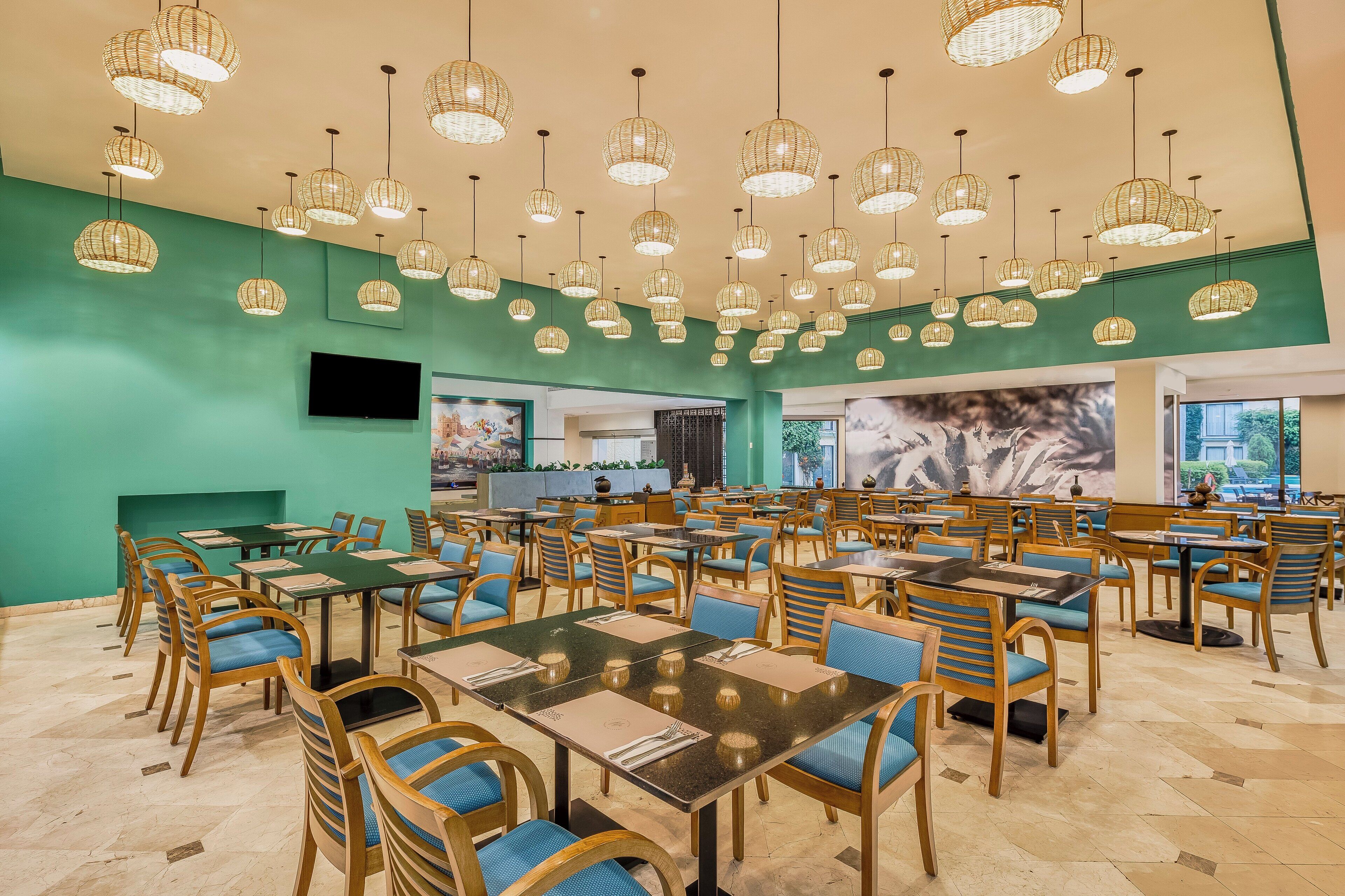 Un vaste espace dédié à la salle à manger, avec de nombreuses tables et chaises, une grande fresque murale et des suspensions en osier.