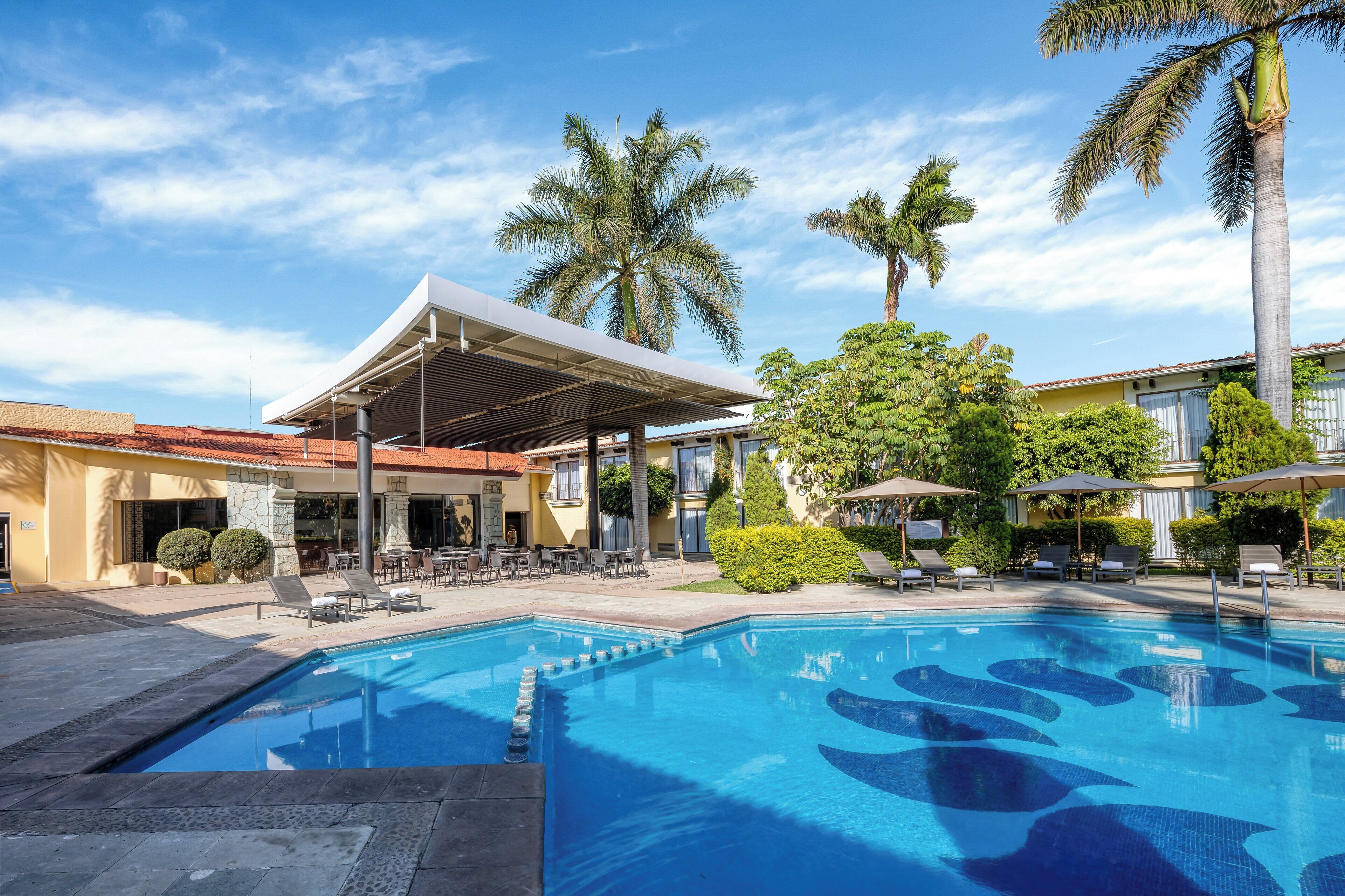 A swimming pool with a modern design, surrounded by lounge chairs and umbrellas, with palm trees and a covered patio area in the background.