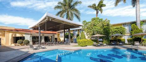 A swimming pool with a modern design, surrounded by lounge chairs and umbrellas, with palm trees and a covered patio area in the background.