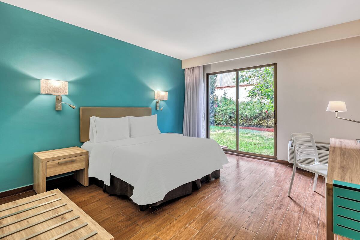 Ein modernes Hotelzimmer mit einem großen Bett, zwei Nachttischlampen, einem hölzernen Nachttisch, einem Schreibtisch mit Stuhl und einem großen Fenster mit Blick auf Grünflächen.