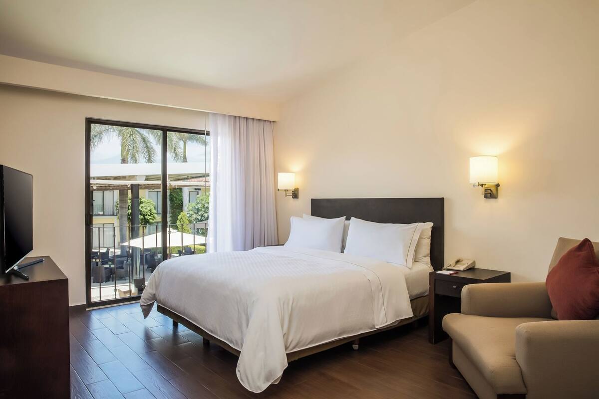 Ein Hotelzimmer mit Bett, Fernseher, Schreibtisch, Stuhl und einem Balkon mit Gartenmöbeln.