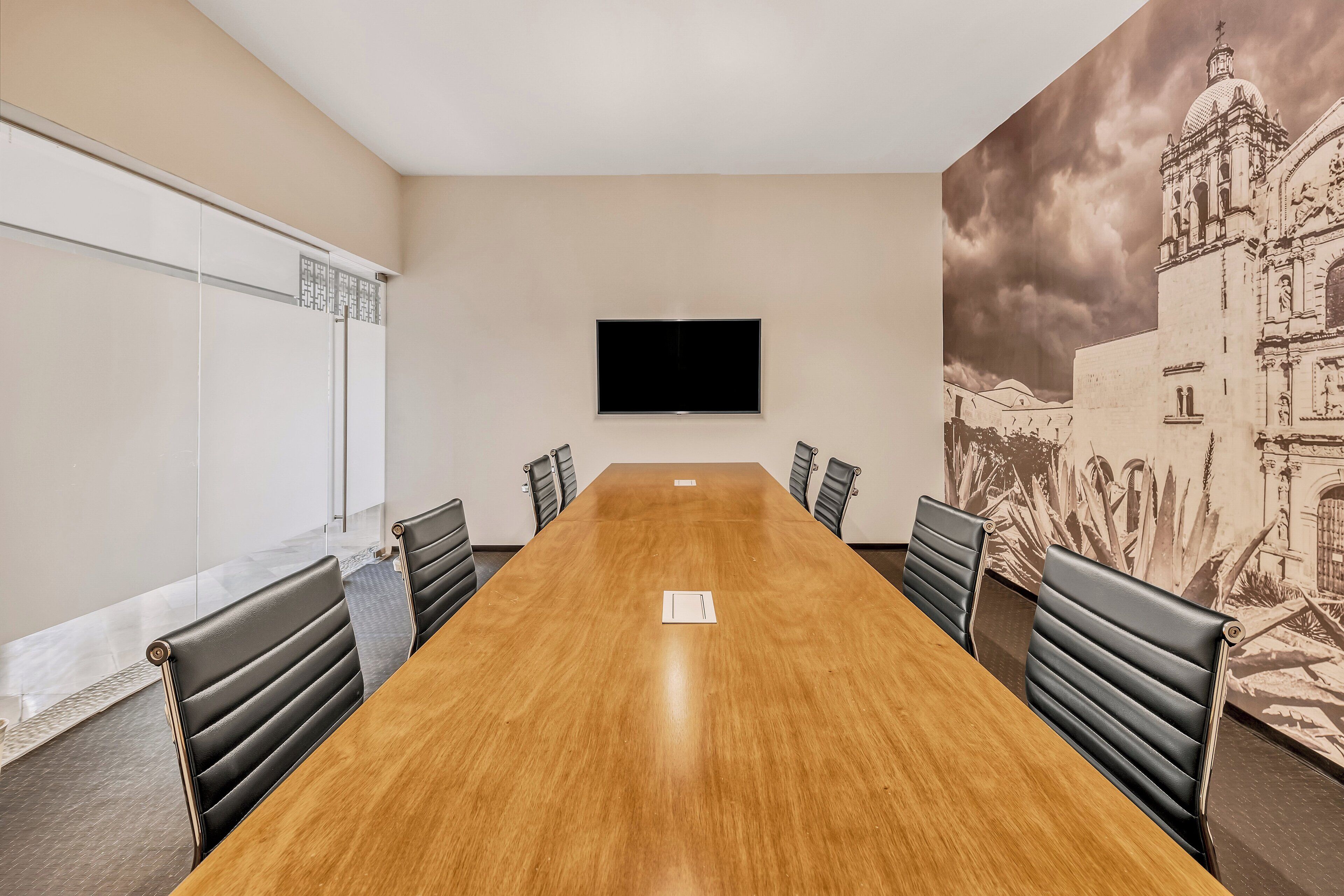 A conference room with a long wooden table, black leather chairs, a flat-screen TV mounted on the wall, and a large mural of a building on the right wall.