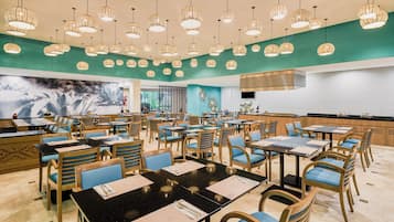 Un comedor amplio con varias mesas y sillas, un mural grande en la pared y una zona de cocina al fondo.