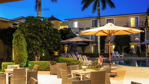 An outdoor seating area with wicker furniture and umbrellas, a pool, and a building with balconies in the background. - Fiesta Inn Oaxaca (Oaxaca)