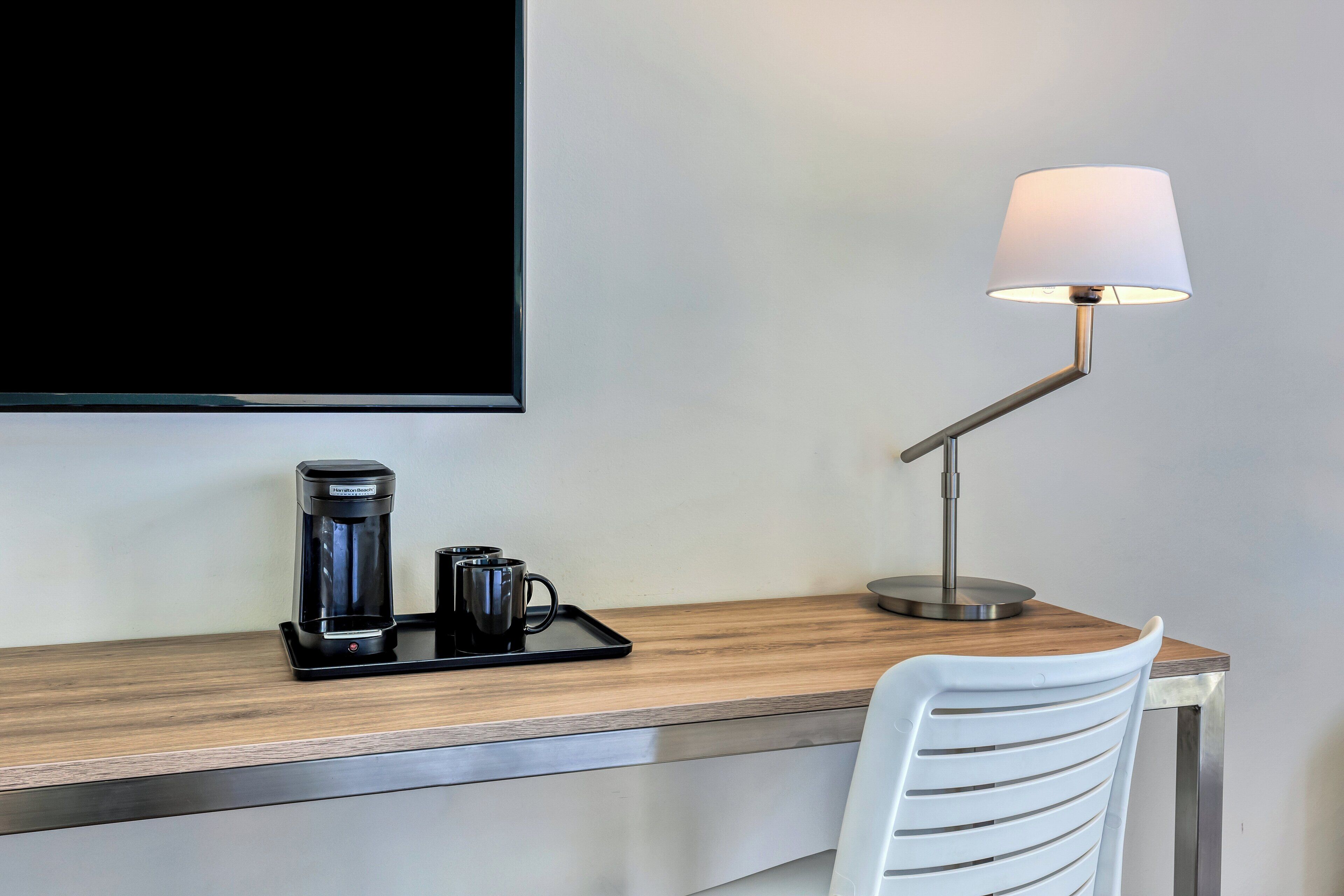 A hotel desk with a coffee maker, two mugs, a tray, and a lamp.
