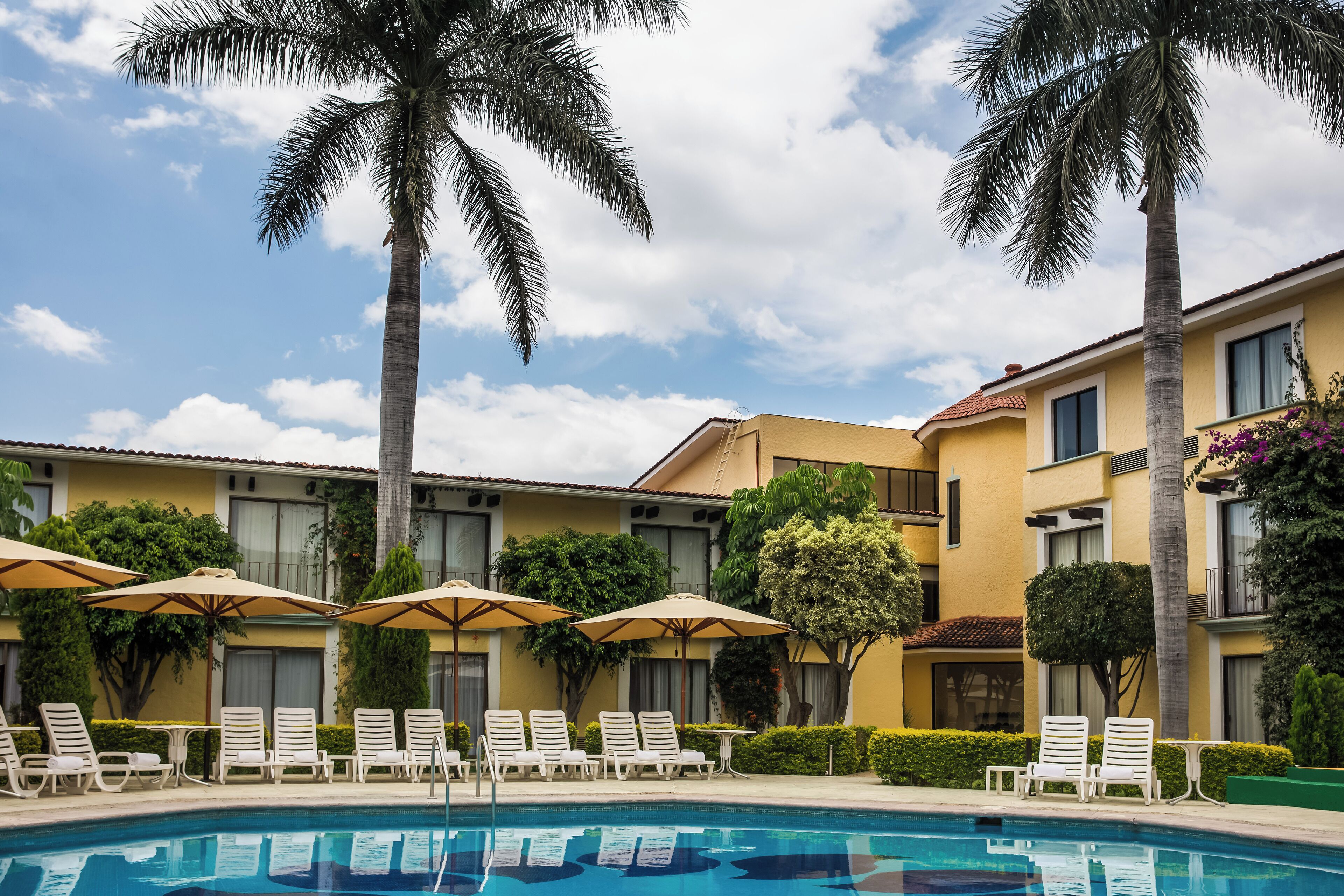 A pool area with lounge chairs and umbrellas, surrounded by palm trees and a building with balconies.
