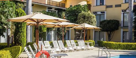 A pool area with lounge chairs and umbrellas, a lifebuoy, and a building with a tiled roof.