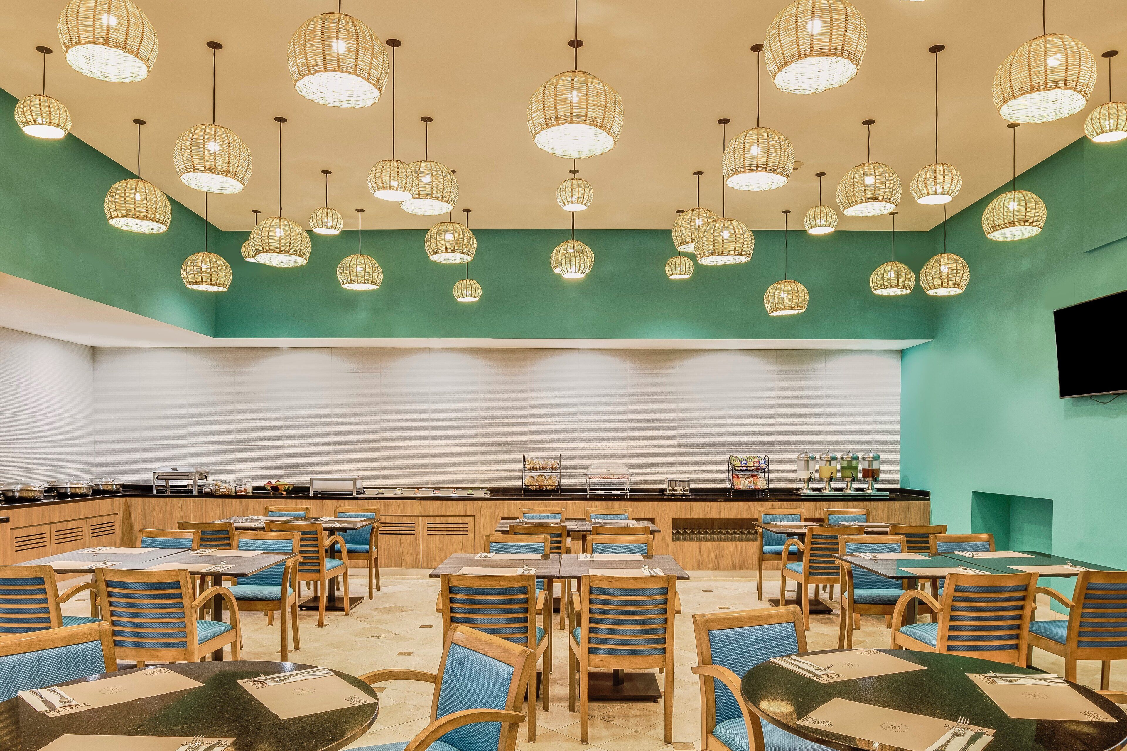 A restaurant with multiple tables and chairs, a counter with a beverage station, and hanging wicker light fixtures.