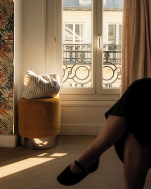 A room with a window, a cushion on a stool, and a person in black attire.