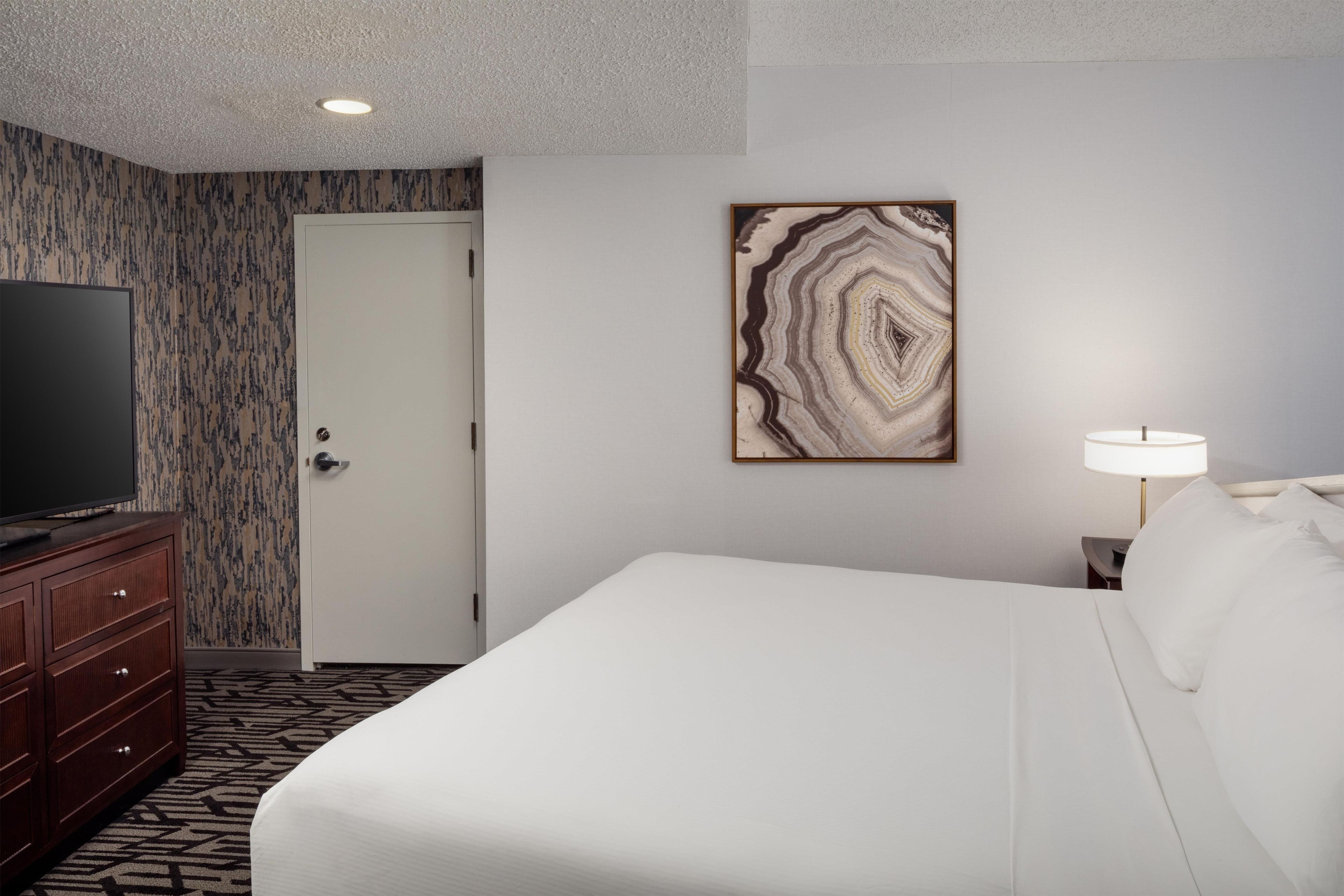 Quarto de hotel com cama, televisão sobre uma cômoda de madeira, porta e uma obra de arte pendurada na parede.