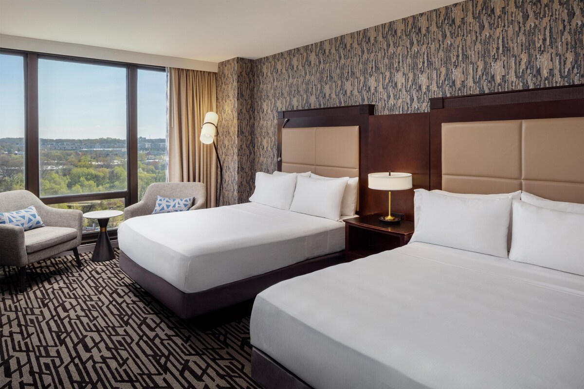 Habitación de hotel con dos camas, una mesita, una silla y vistas a la ciudad.