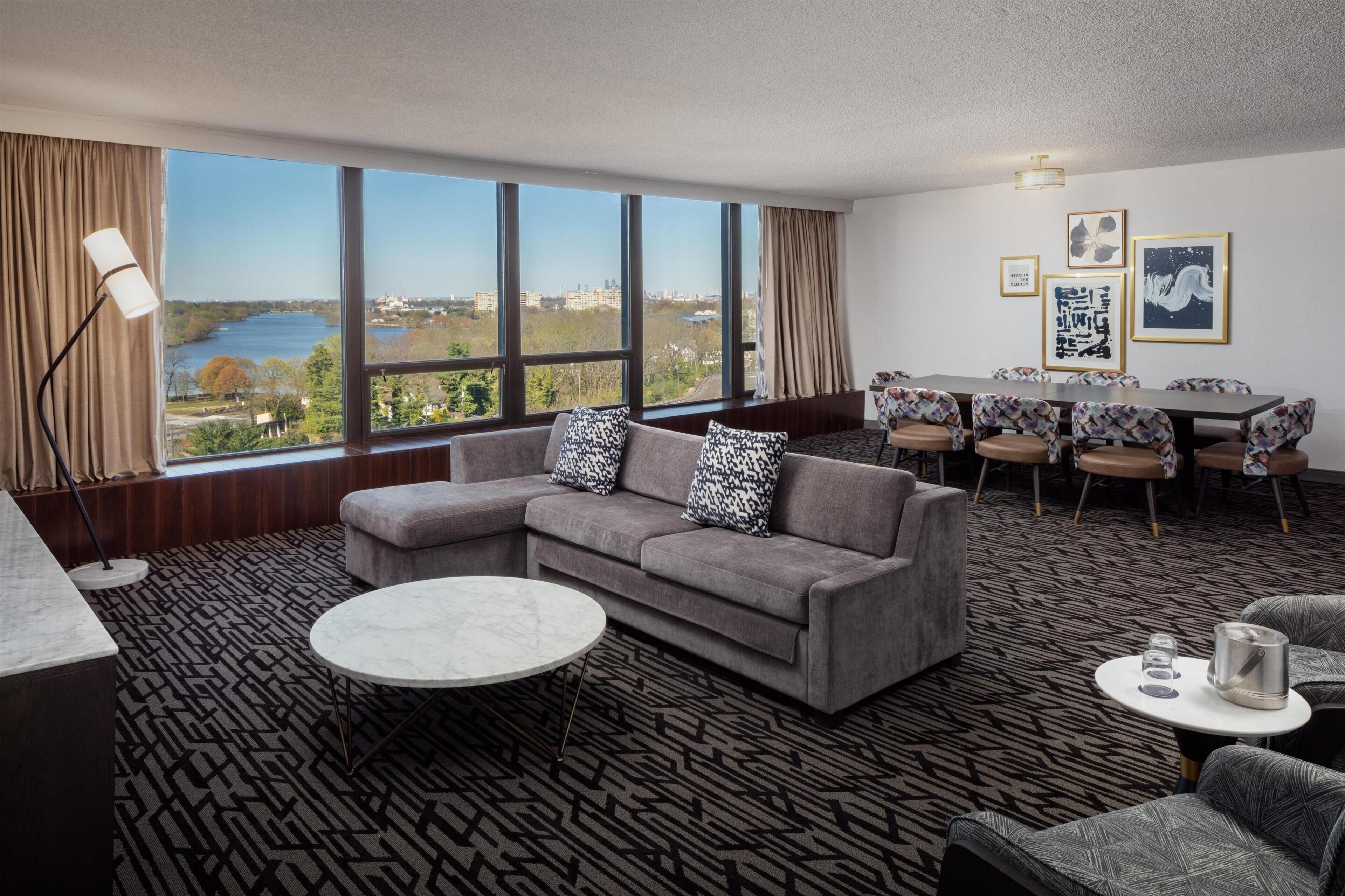 A modern hotel room with a large window, a gray sofa, a round coffee table, and a dining area with patterned chairs.