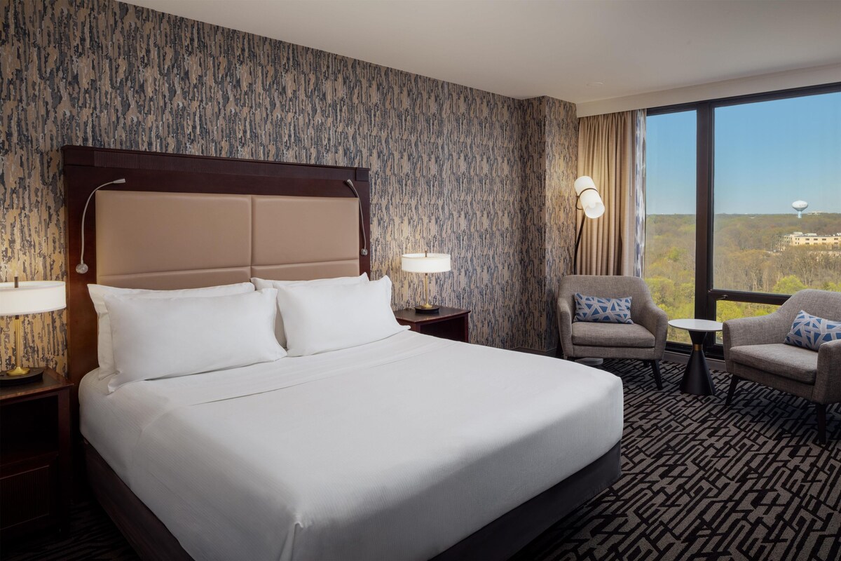 Habitación de hotel con una cama grande, dos sillones, una mesita y vistas a un paisaje urbano a través de amplios ventanales.