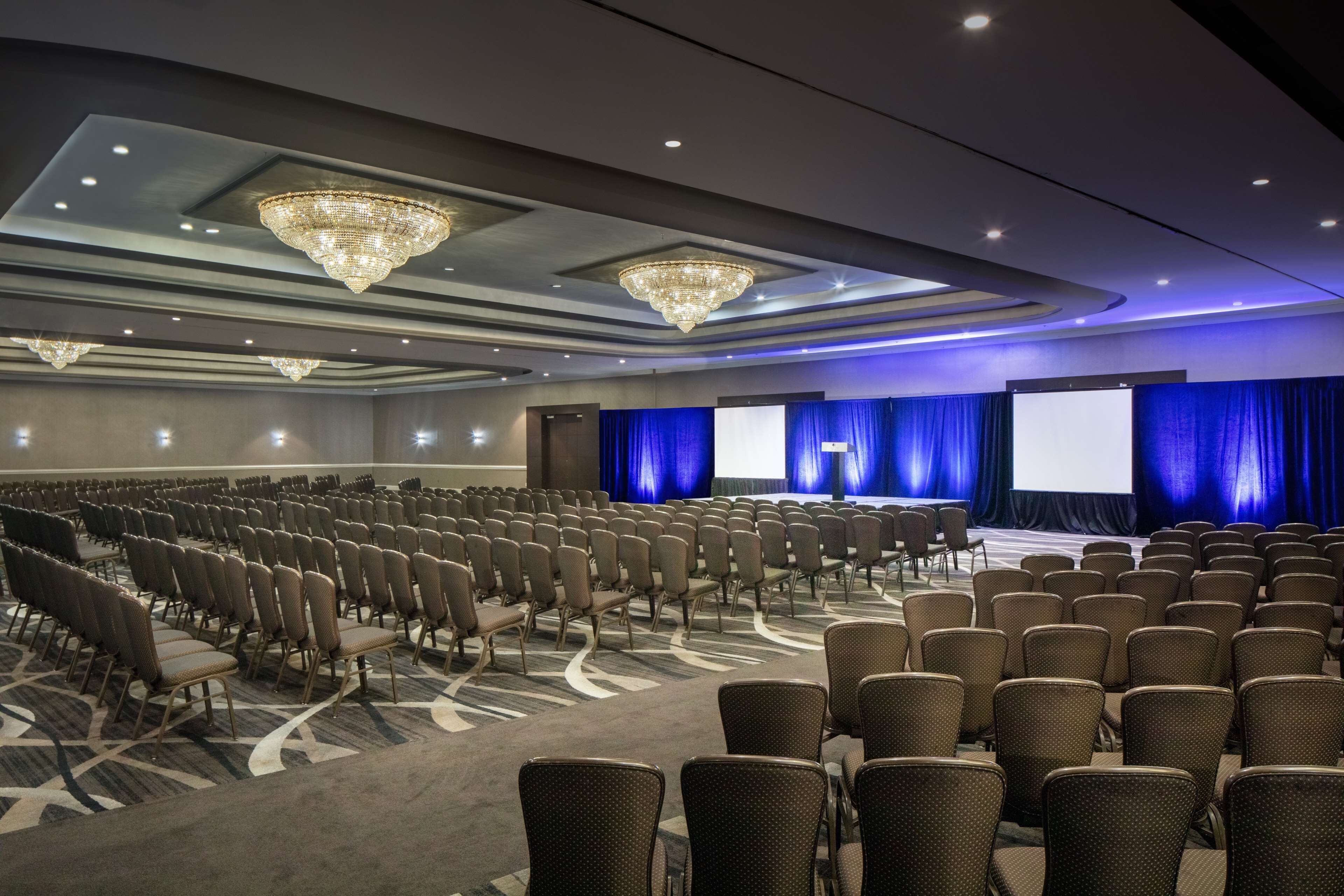 A conference room with rows of chairs, a stage with a screen, and chandeliers.