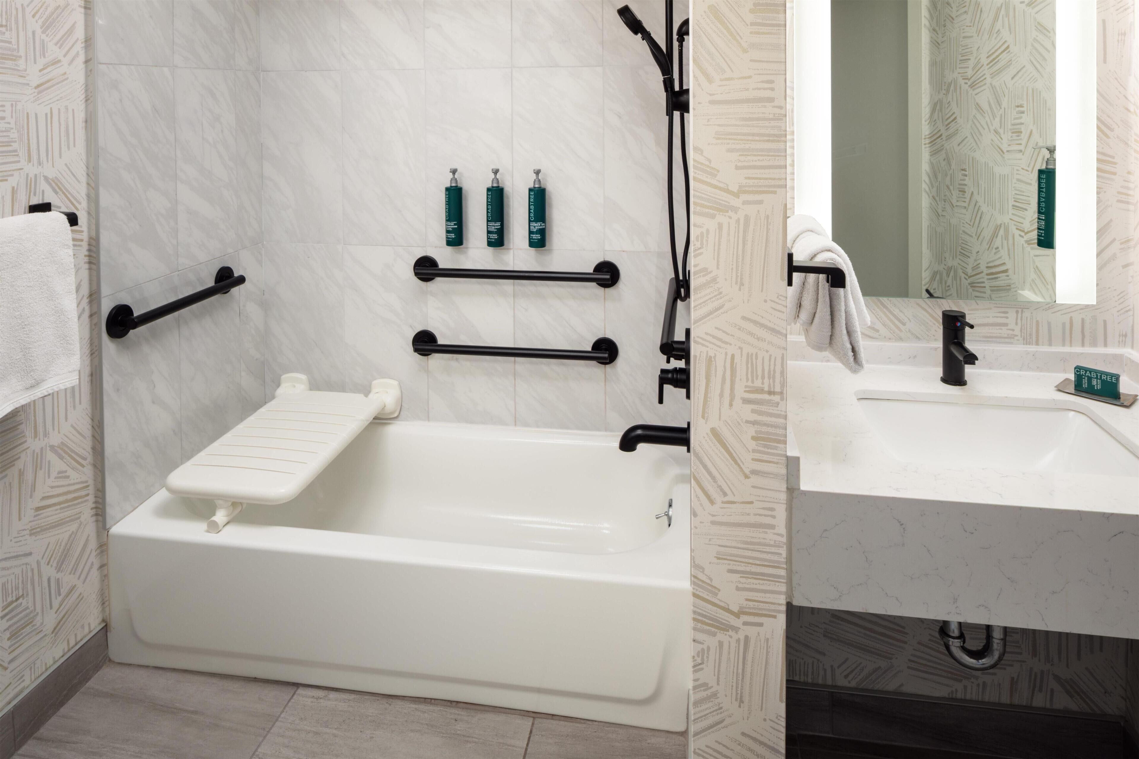 Un baño moderno con bañera, lavamanos y espejo. Hay barras de apoyo, un perchero para toallas y artículos de aseo en la pared.