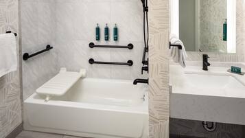 Un baño moderno con bañera, lavamanos y espejo. Hay barras de apoyo, un perchero para toallas y artículos de aseo en la pared.