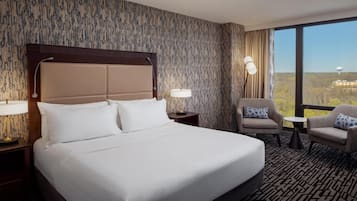 Habitación de hotel con una cama grande, dos sillones, una mesita y vistas a un paisaje urbano a través de amplios ventanales.