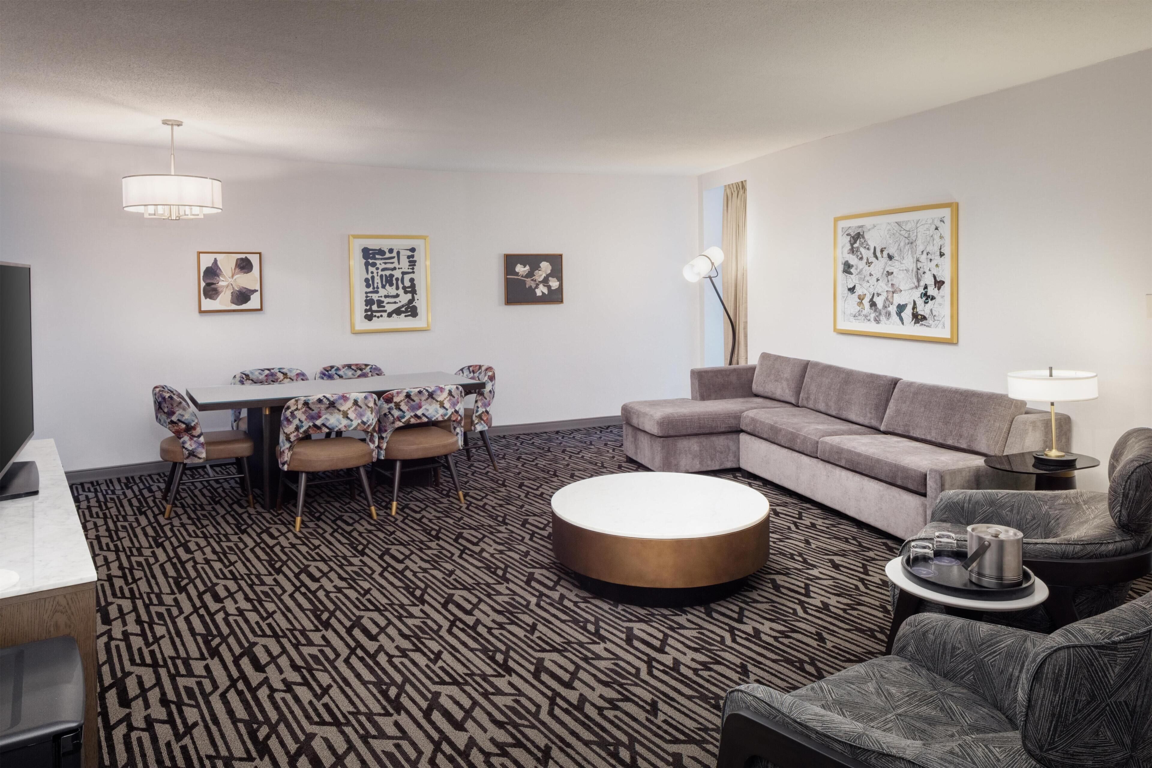 Una habitación de hotel moderna con comedor, un sofá, una mesa de centro redonda y obras de arte en las paredes.
