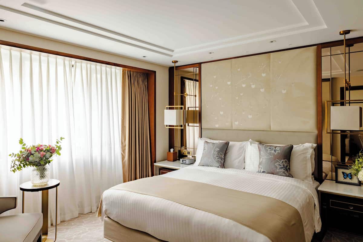 A hotel room with a large bed, a bedside table, a nightstand, a mirror, and a window with curtains.
