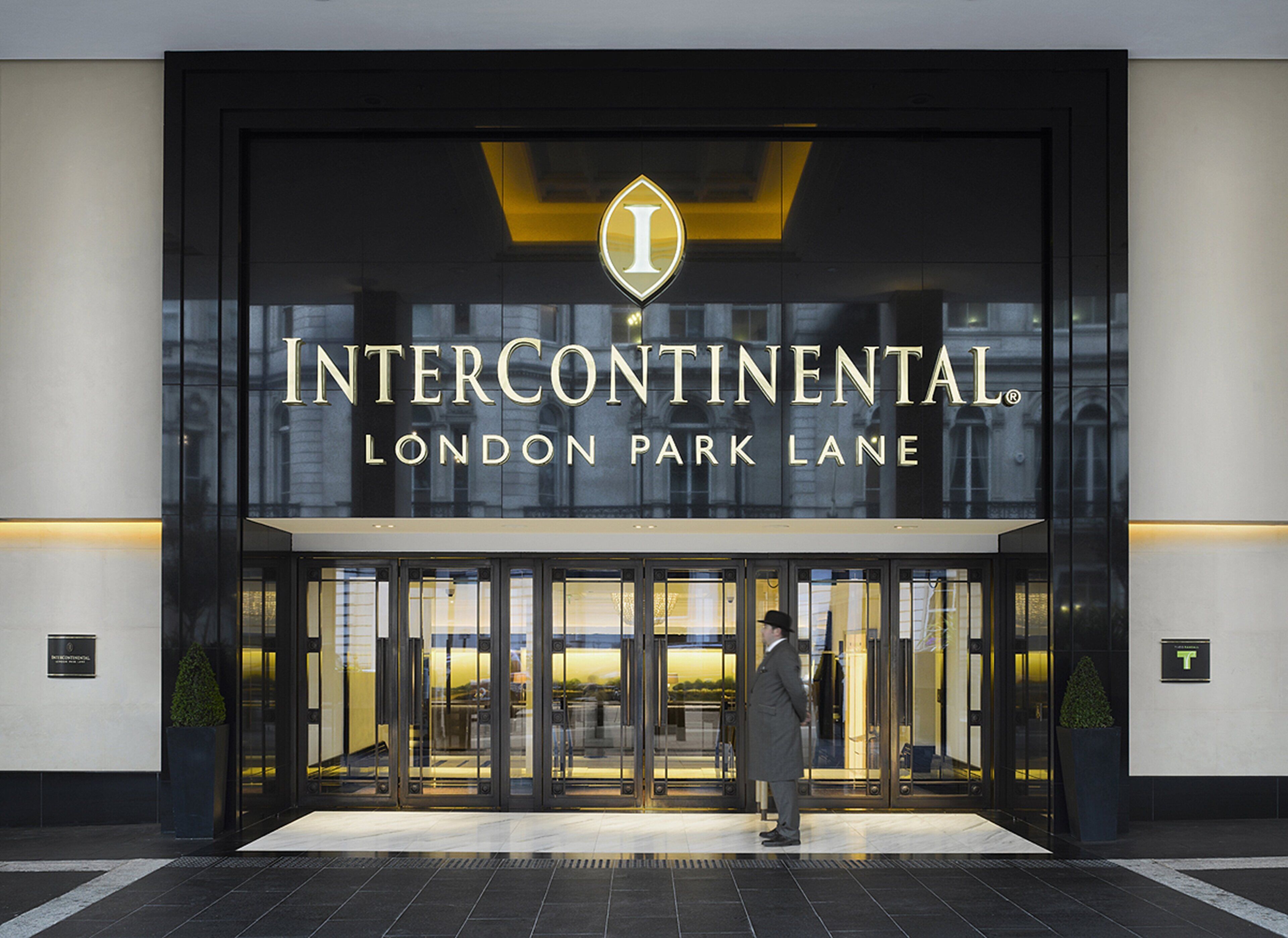 The entrance to Intercontinental London Park Lane hotel with a glass facade and a man standing in front.