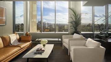 Ein modernes Wohnzimmer mit einem Ledersofa, einem weißen Sessel und einem Couchtisch mit Büchern und einer Vase. Große Fenster bieten Blick auf die Stadt.