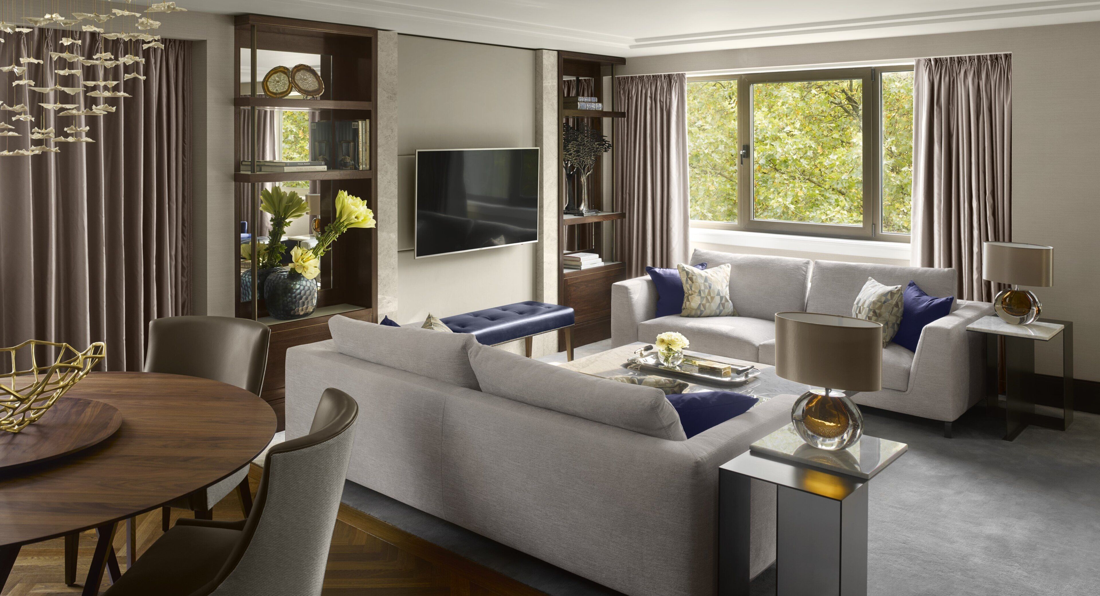 A modern living room with a sofa, armchairs, a coffee table, and a TV. There is a bookshelf and a window with curtains.