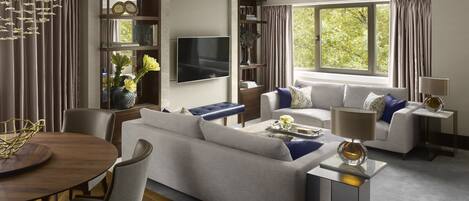 A modern living room with a sofa, armchairs, a coffee table, and a TV. There is a bookshelf and a window with curtains.
