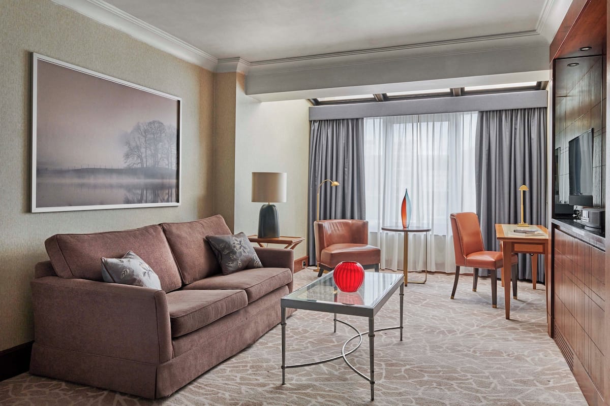 A modern hotel room with a brown sofa, a glass coffee table, a TV, and a large window with curtains.
