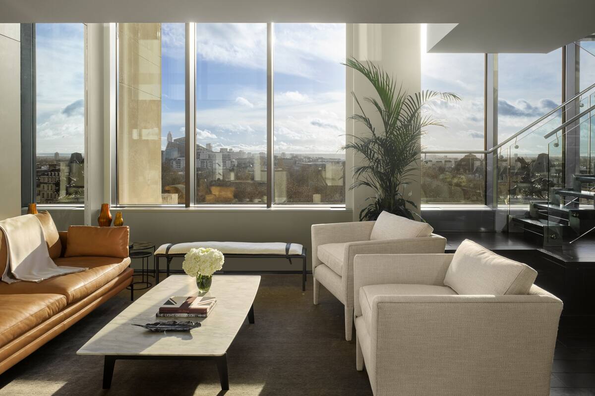 A modern living room with a leather sofa, a white armchair, and a coffee table with books and a vase of flowers. Large windows offer a city view.