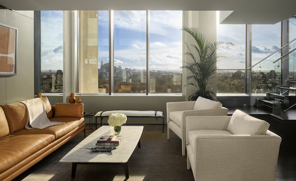 Una sala de estar moderna con un sofá de cuero, un sillón blanco y una mesa de centro con libros y un jarrón de flores. Amplias ventanas ofrecen vistas de la ciudad.