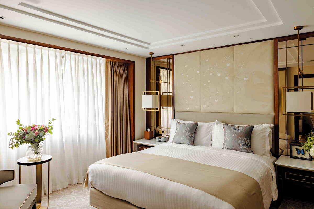 A hotel room with a large bed, a bedside table, a nightstand, a mirror, and a window with curtains.