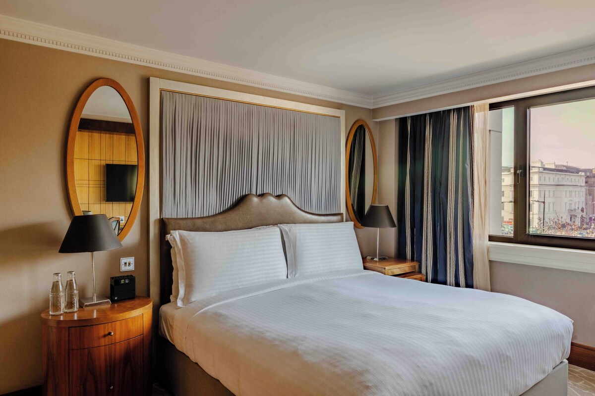 A hotel room with a large bed, a wooden nightstand with a lamp, a mirror, and a view of the city through a window.