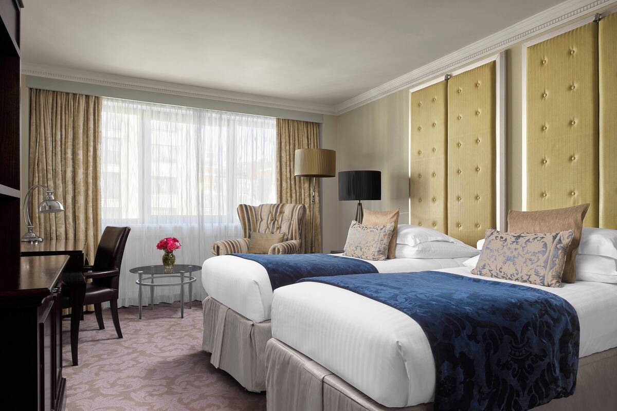 A hotel room with two beds, a desk, a chair, a lamp, and a window with curtains.