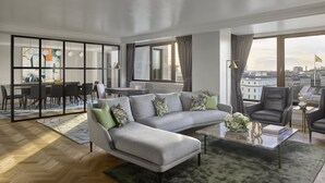 A modern living room with a grey sectional sofa, a coffee table, and two leather armchairs. There is a dining area with a table and chairs, and large windows offering a city view. - InterContinental London Park Lane by IHG (London)