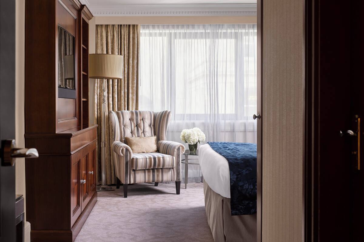 A hotel room with a large wooden wardrobe, a striped armchair, a bed with a blue blanket, and a window with sheer curtains.