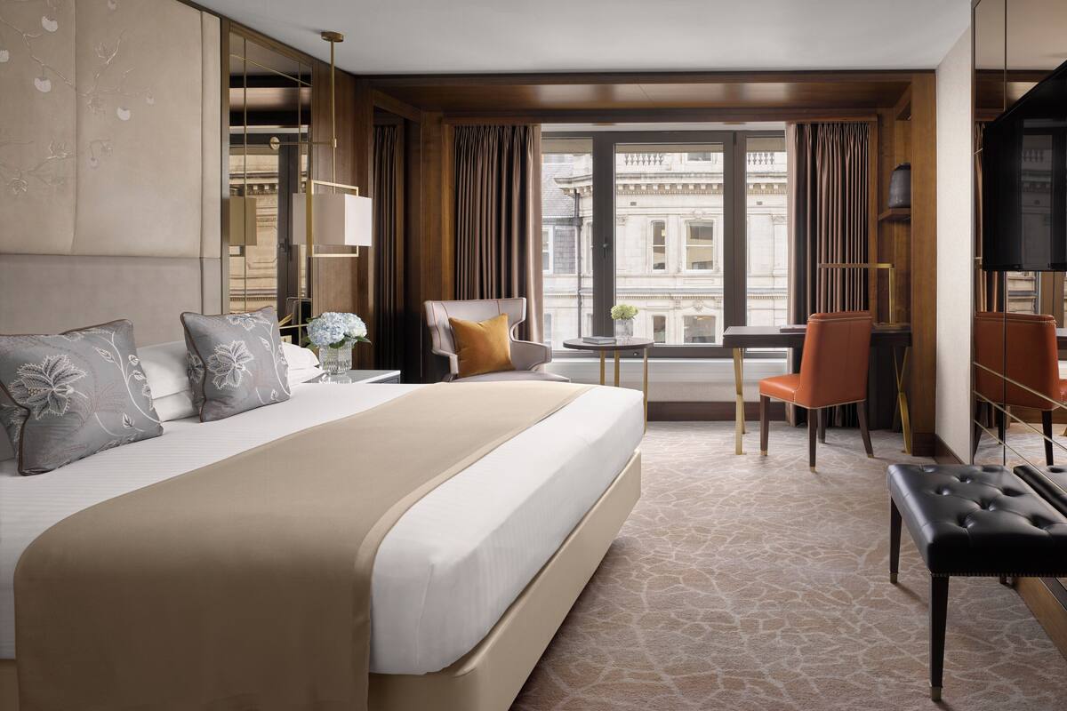 A hotel room with a large bed, a desk with two chairs, a television, and a view of the city through large windows.