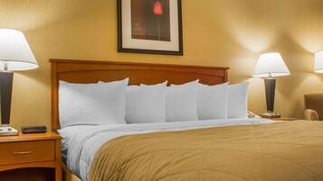 Habitación de hotel con cabecera de madera, cama con ropa blanca, colcha marrón, dos lámparas de noche, mesita de noche con teléfono e imagen enmarcadas en la pared.