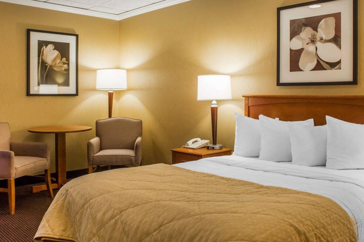 A hotel room with a large bed, two chairs, a small table, and two lamps.