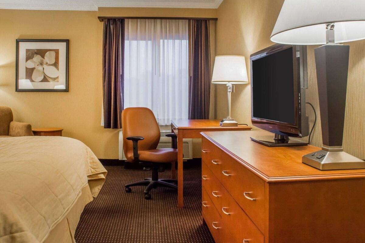 A hotel room with a bed, a desk with a television, a chair, a dresser, and a window with curtains.
