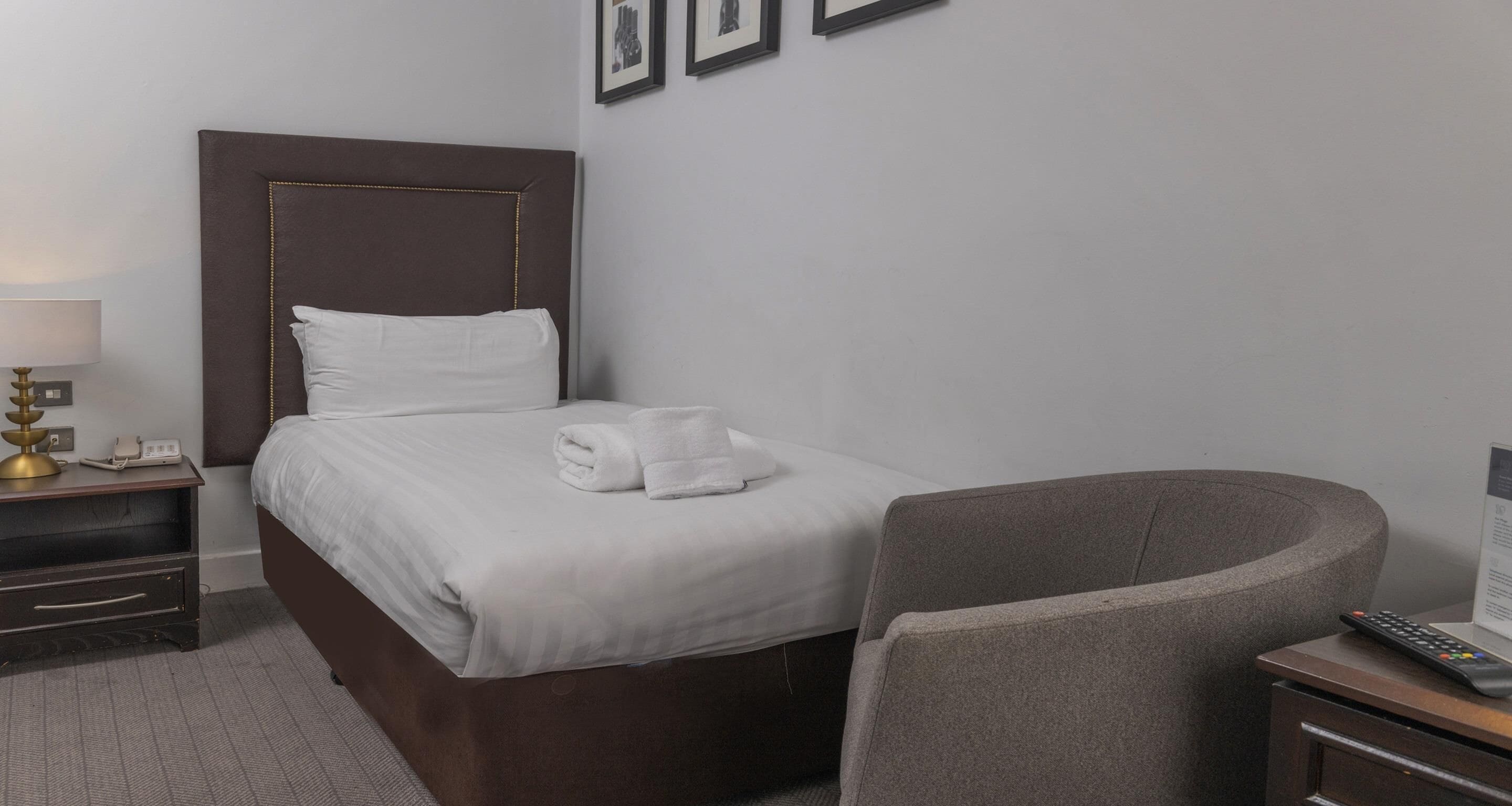 A hotel room with a bed, a nightstand, a lamp, a chair, and a remote control.