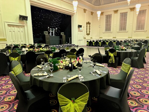 A banquet hall with round tables set for a formal event, featuring black chairs with green sashes, and a stage with a dark backdrop. - Carlisle Station Hotel, Sure Hotel Collection by BW (Carlisle)