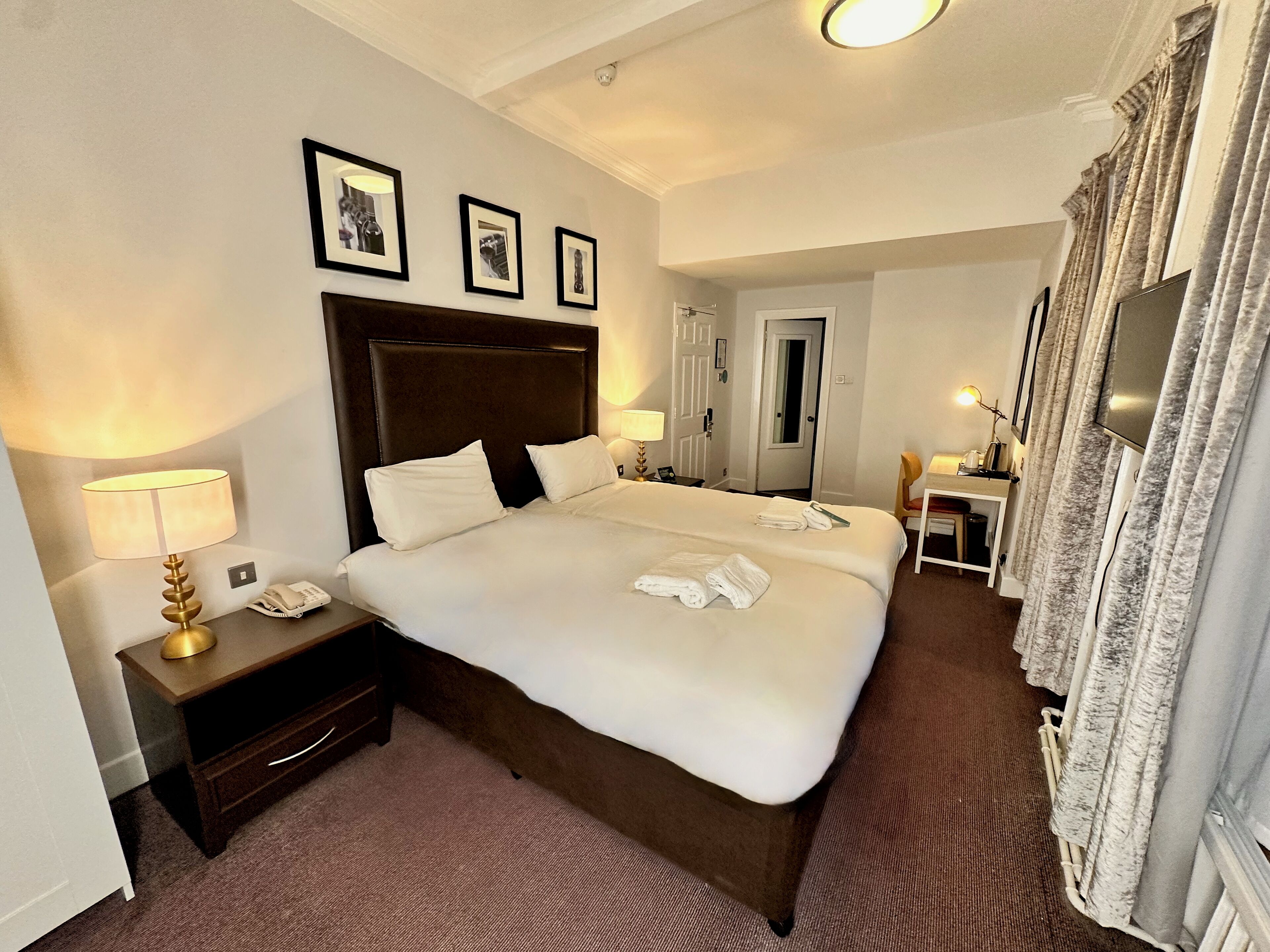 Habitación de hotel con una cama grande, una mesita de noche con lámpara, un escritorio con silla y una televisión.
