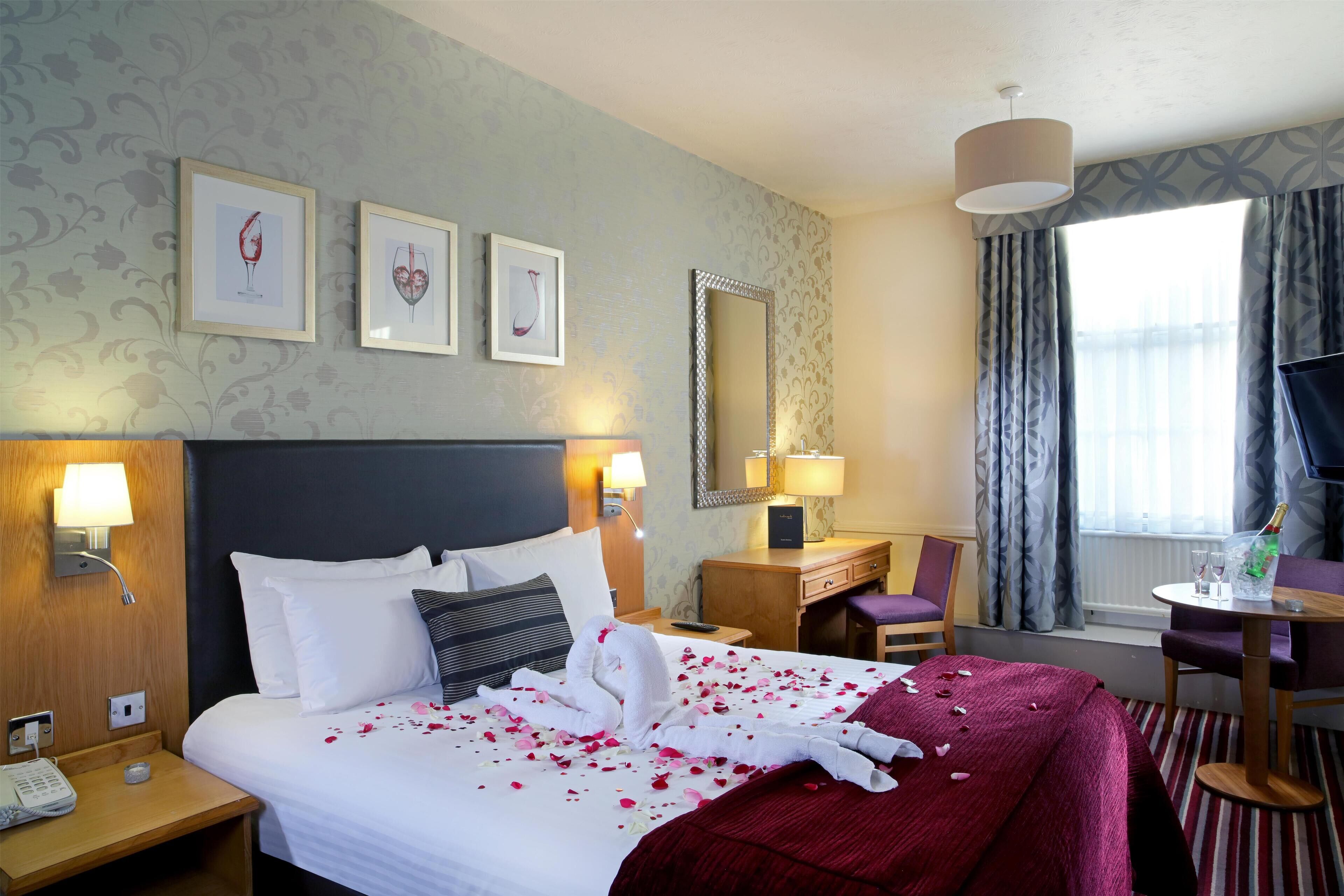 A hotel room with a bed, bedside table, lamp, mirror, desk, chair, and a window with curtains.