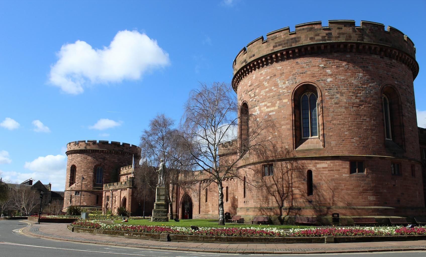 A historic building with two round towers and arched windows, surrounded by a landscaped garden with flowers and benches.