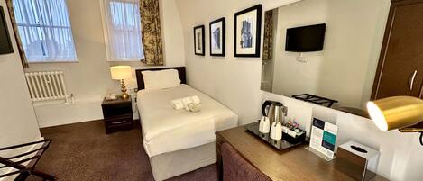 Habitación de hotel con cama, mesita de noche, lámpara, cafetera y escritorio con una silla.