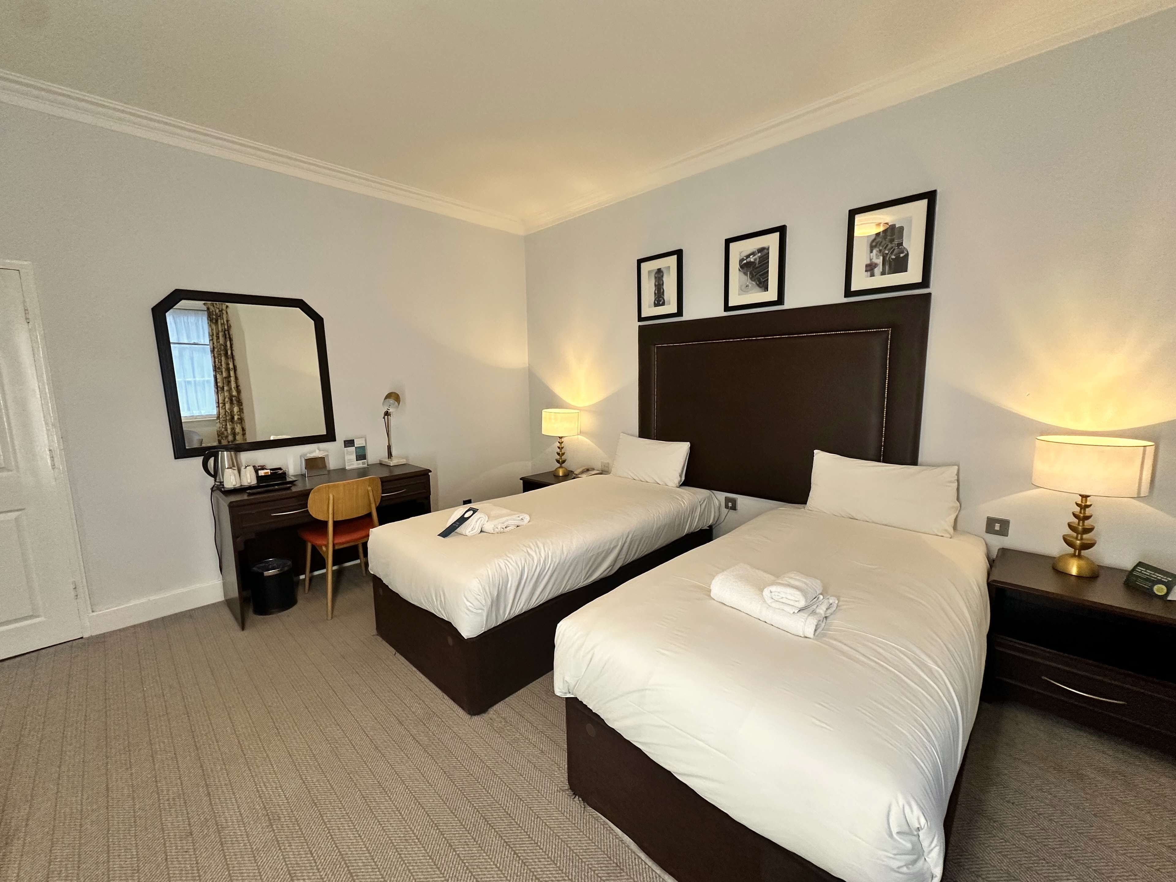 Habitación de hotel con dos camas, un escritorio, una silla, un espejo y lámparas de noche.