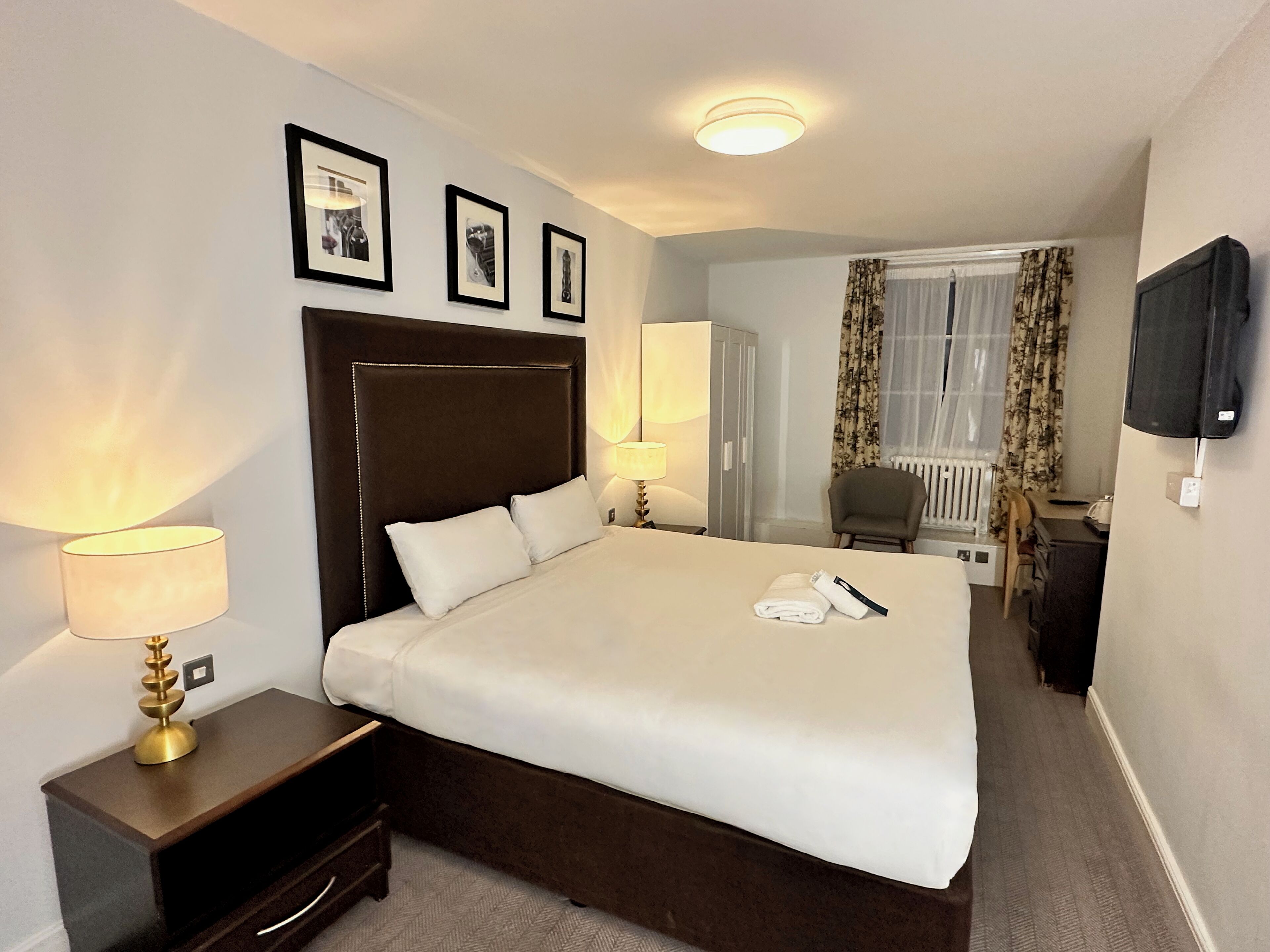 A hotel room with a large bed, two bedside lamps, a flat-screen TV mounted on the wall, and a window with curtains.